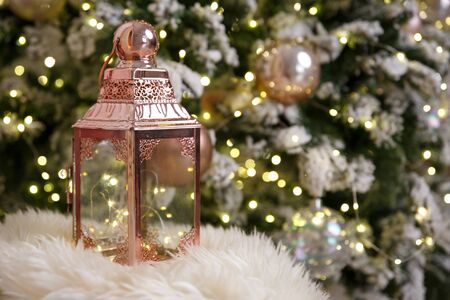 Christmas card and background. A pink lantern stands on a white fur plaid, against a background of bokeh of yellow lights of a Christmas tree garlandの写真素材