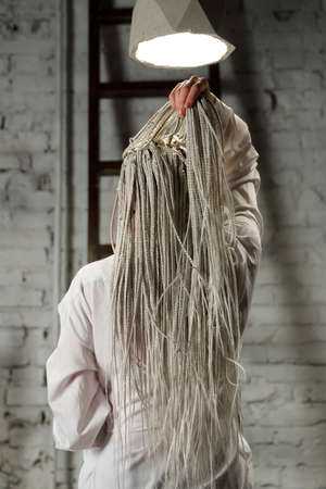 Woman with white braids, in shirt. Young woman sits with her back under the light of concrete chandeliers.の写真素材
