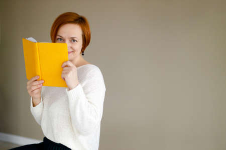 woman covered her face with diary. Business red haired woman in white sweater holding notebook in her hand and smiling. Business planning and tasks for the day or week. Female in businessの写真素材