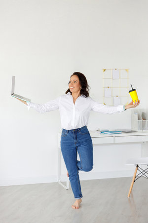 business woman stands on one leg in balance pose, holding laptop and coffee. Concept: choice between work and rest. Stress and deadline, motivation. Hard worker femaleの写真素材