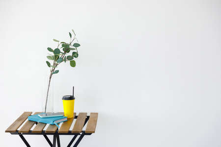 Wooden table against background of white wall. Vase with eucalyptus branch, yellow paper coffee cup...