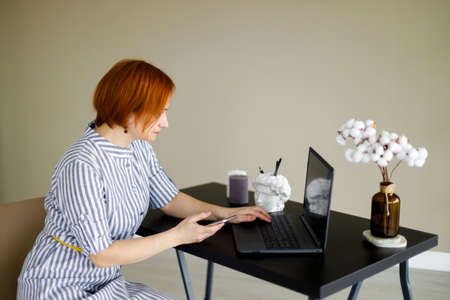 red haired woman in striped dress sits against background of beige wall, works at a laptop. Distant work. Online training. Home officeの写真素材