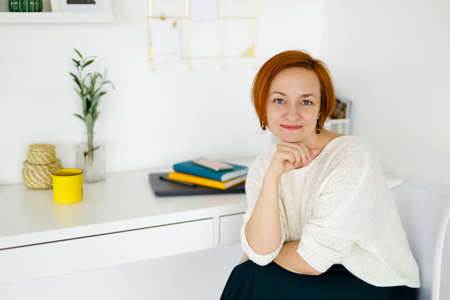 red haired woman in a white sweater on a light background smiling and looking at the camera. Distant...
