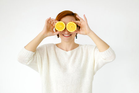 red haired woman smiles with snow white smile and covered her eyes with lemons. Strange face. Grimace. Woman in white sweater on white background, isolated. Dental health, fresh breathの写真素材