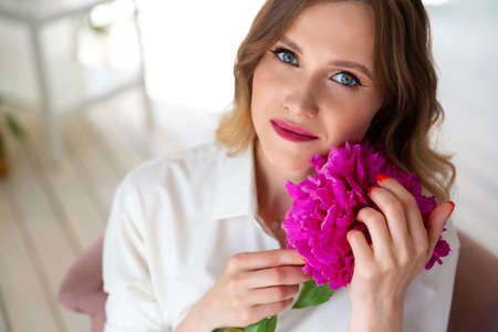 woman in shirt, holds bouquet of peonies in her hands, looks at camera. Portrait of beautiful blonde woman. Surprise bouquet of flowers for date, mothers day or valentines dayの写真素材