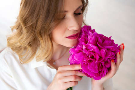 woman in white shirt, holding peony bud in her hands, closed her eyes. Portrait of beautiful blonde woman. Surprise bouquet of flowers for date, mothers day or valentines day.の写真素材