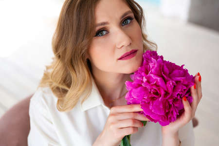 woman in shirt, holding peony bud in her hands, looks at camera. Portrait of beautiful blonde woman. Surprise bouquet of flowers for date, mothers day or valentines dayの写真素材