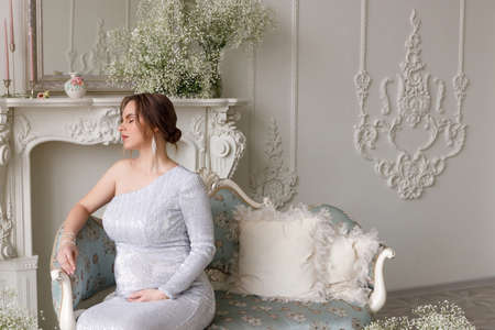 portrait of beautiful pregnant woman in silver evening dress with sequins sitting on sofa in white...