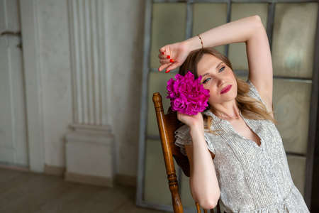woman in white dress, holding peony bud in her hands, looking away. Portrait of beautiful blonde woman. Surprise bouquet of flowers for date, mothers day or valentines day. Place for your text.の写真素材