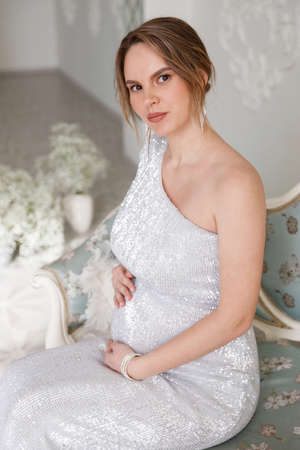 beautiful pregnant woman in evening dress sits on a sofa in a white room. The concept of motherhood and the birth of children. Glamor and beautiful lifeの写真素材