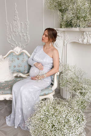 beautiful pregnant woman in evening dress sits on a sofa in a white room. The concept of motherhood and the birth of children. Glamor and beautiful life. Room decorated with gypsophila flowersの写真素材