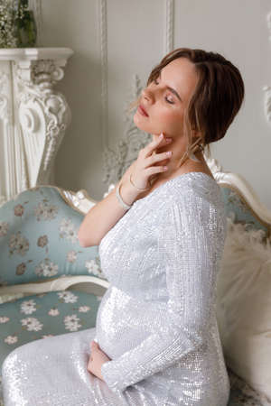 beautiful pregnant woman in evening dress sits on a sofa in a white room. The concept of motherhood and the birth of children. Glamor and beautiful lifeの写真素材