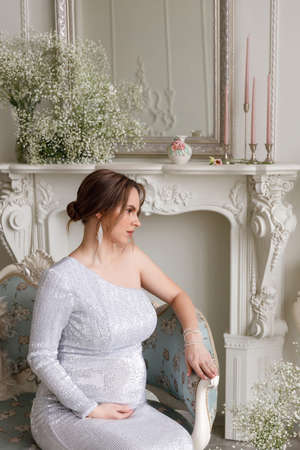 portrait of beautiful pregnant woman in silver evening dress with sequins sitting on sofa in white room. The concept of motherhood, parenthood and the birth of children. Glamor and beautiful lifeの写真素材