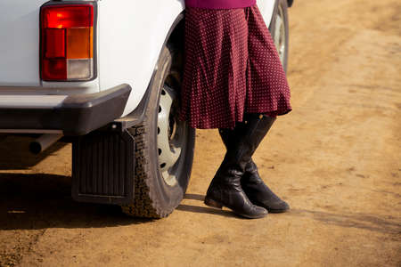 cropped image of female legs. A woman in a sweater, skirt and boots stands on the sand near the car