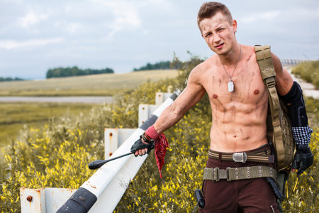 male vandal with dirty naked torso and mace in his hand, against backdrop of an overgrown abandoned roadの写真素材