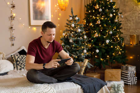 brunet man in T-shirt and jeans sits on bed in room decorated for Christmas, holding smartphone in his hands. Communication on social networks. Leisure and work, freelanceの写真素材