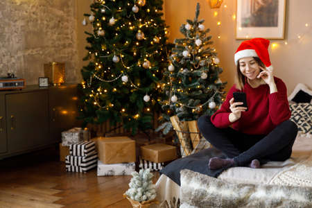 blonde woman in red sweater and jeans sits on bed in room decorated for Christmas, holding...
