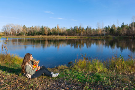 a woman with a small dog yorkshire terrier sits on the grass on the bank of a pond in the parkの写真素材