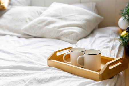 wooden tray and two white mugs on the bed on a white sheet, near a decorated Christmas tree. Cozy...