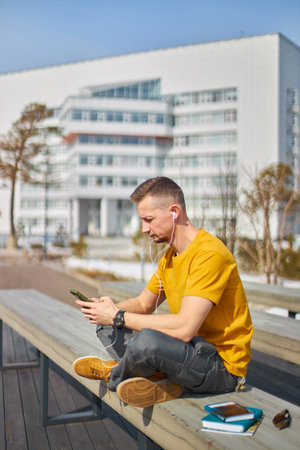 man student with a smartphone in his hands and headphones, against the backdrop of the university buildingの写真素材