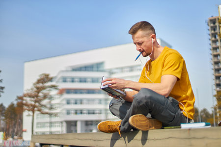 man student with a smartphone in his hands and headphones, against the backdrop of the university buildingの写真素材