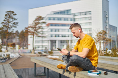 man student with a smartphone in his hands and headphones, against the backdrop of the university buildingの写真素材