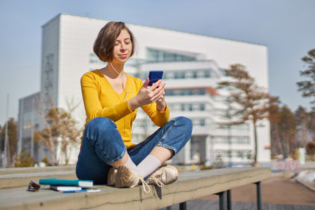 woman student with a smartphone in her hands and headphones, against the backdrop of the university buildingの写真素材