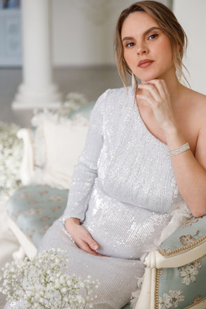 beautiful pregnant woman in evening dress sits on a sofa in a white room. The concept of motherhood and the birth of children. Glamor and beautiful lifeの写真素材