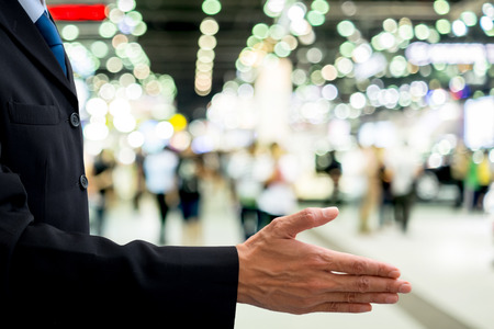 Isolated businessman in dark gray suit offer his right hand for handshake for global business, cooperation and partnership. Defocus exhibition in the backgroundの写真素材