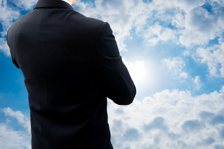 Back side of businessman in dark gray suit standing and crossing one's arm watching world economy situation and thinking about global business in summer cloudy sky backgroundの写真素材