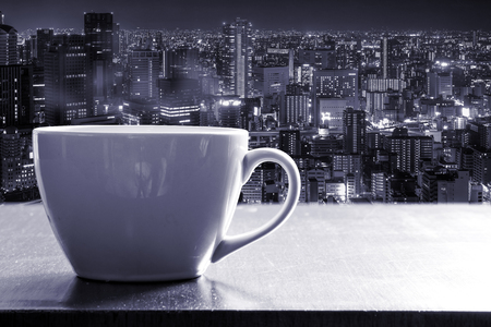 Hot coffee in white cup on wooden counter bar beside the glass window on high tower with Osaka city night view. Monotone effect.の写真素材