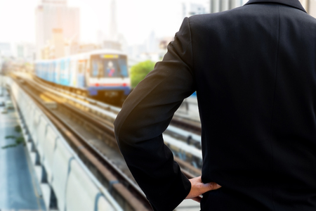 Back side of businessman in dark gray suit standing and akimbo waiting for the train in railway...
