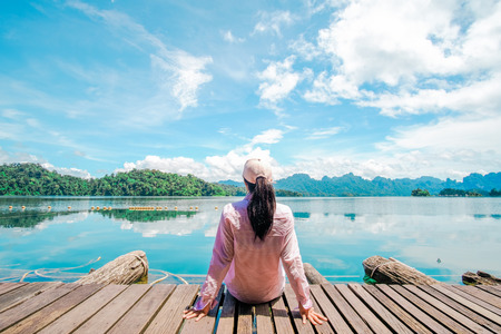 Woman sitting on the wooden floor watching out to the lake, forest and blue sky in summer.の写真素材