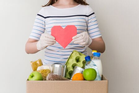 A girl in medical gloves dial charity donation cardboard box with food: oil, fruits, cereals, canned food. White background, table in the kitchen. Pink heart postcardの写真素材