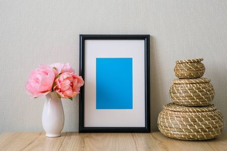 Mockup of a black vertical photo frame with Passepartout. The light interior in the Scandinavian style. Composition on the table, shelf. Pink peonies in a white vase, wicker round basketsの写真素材