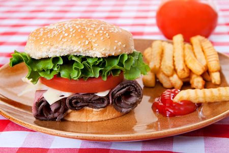 one large roast beef sandwich in sesame roll with tomatos and lettuce on plate with classic red and white checkered tableclothの写真素材