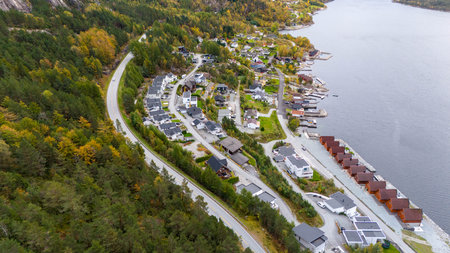 Aerial view of a scenic village near a fjord with modern houses, surrounded by lush green forests and a winding road. The fjord is calm, and the houses are neatly arranged along the shoreline.の写真素材