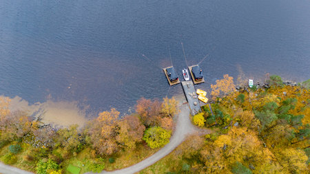 Aerial view of a serene lake with a wooden dock. Several boats are moored at the dock, two of which...