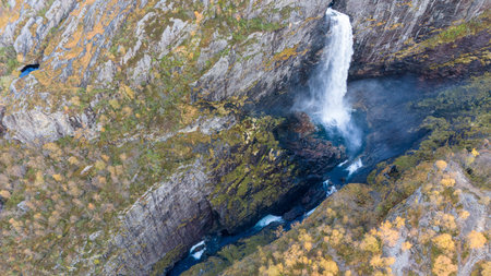 Aerial view of a stunning waterfall cascading down rocky cliffs into a deep blue pool surrounded by lush greenery and autumn foliage.の写真素材