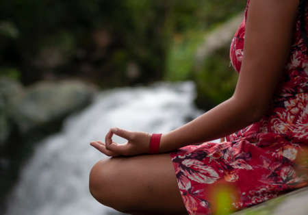woman in red dress meditating in front of a waterfall, her hand is in yoga position and she has a...