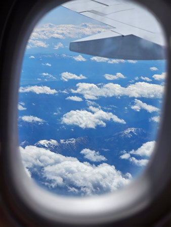 Snow top Alps Mountains seen from airplane window mid-flight through scattered white clouds on a...