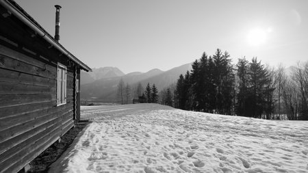 Wooden hut with shutters and narrow chimney in the snow where footsteps can be seen and sunrays, forest and mountains in background in black and white in Austriaの写真素材