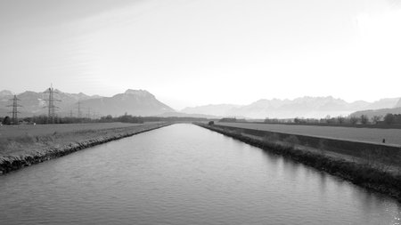The Rhine River on a summer day between Austria and Switzerland with bicyclist on the dam path and power poles and mountains in the background in black and whiteの写真素材