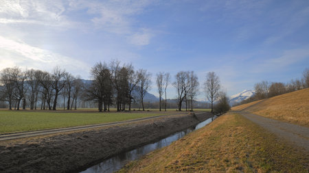 Dam road next to river in winter with bare trees and snow covered mountains in backgroundの写真素材