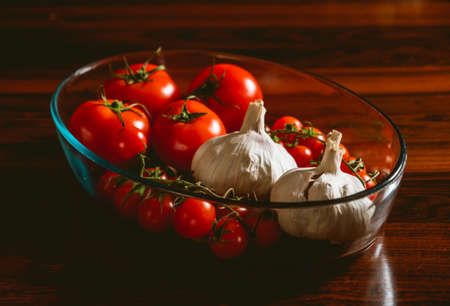 Red tomatoes and garlic heads in a glass bowl over a dark brown wooden table.の写真素材