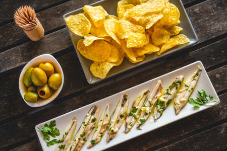 Three dishes with potato chips, picked anchovies and green olives on a dark wooden table with toothpicks. Typical Spanish snacks to eat in a bar.の写真素材