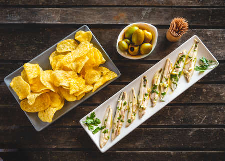 Three dishes with potato chips, picked anchovies and green olives on a dark wooden table with toothpicks. Typical Spanish snacks to eat in a bar.の写真素材