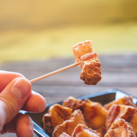 Hand holding a piece of fried pork rinds called "Torreznos" with a toothpick on a dark wooden table. Typicall spanish snacks to eat in a bar. Photo with selective focus.の写真素材