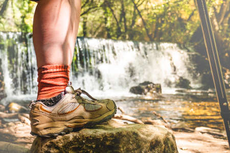 Woman leg with hiking boots and stick on a rock with a natural waterfall behind.の写真素材