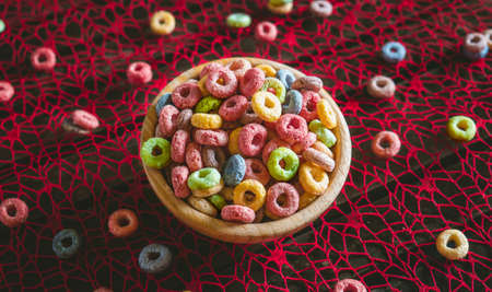 Colorful round fruit cereals in a wooden bowl on a red table, with some scattered outside the bowl....
